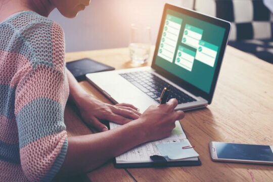 Woman writing in notebook on a table surrounded by laptop and cell phone