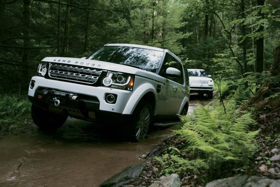 Two white Land Rovers driving through a muddy forest.