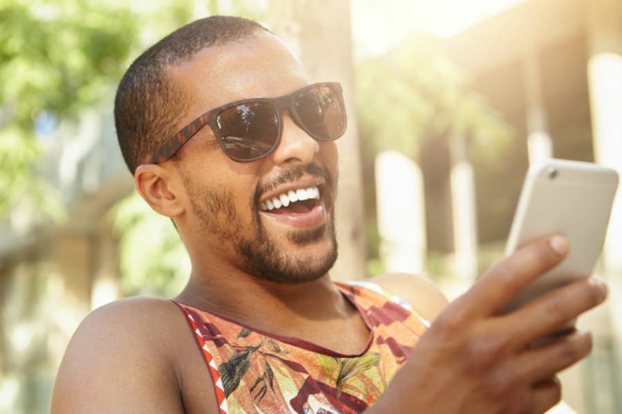 Black male traveler wearing tropical shirt sitting outside looking at smartphone and laughing