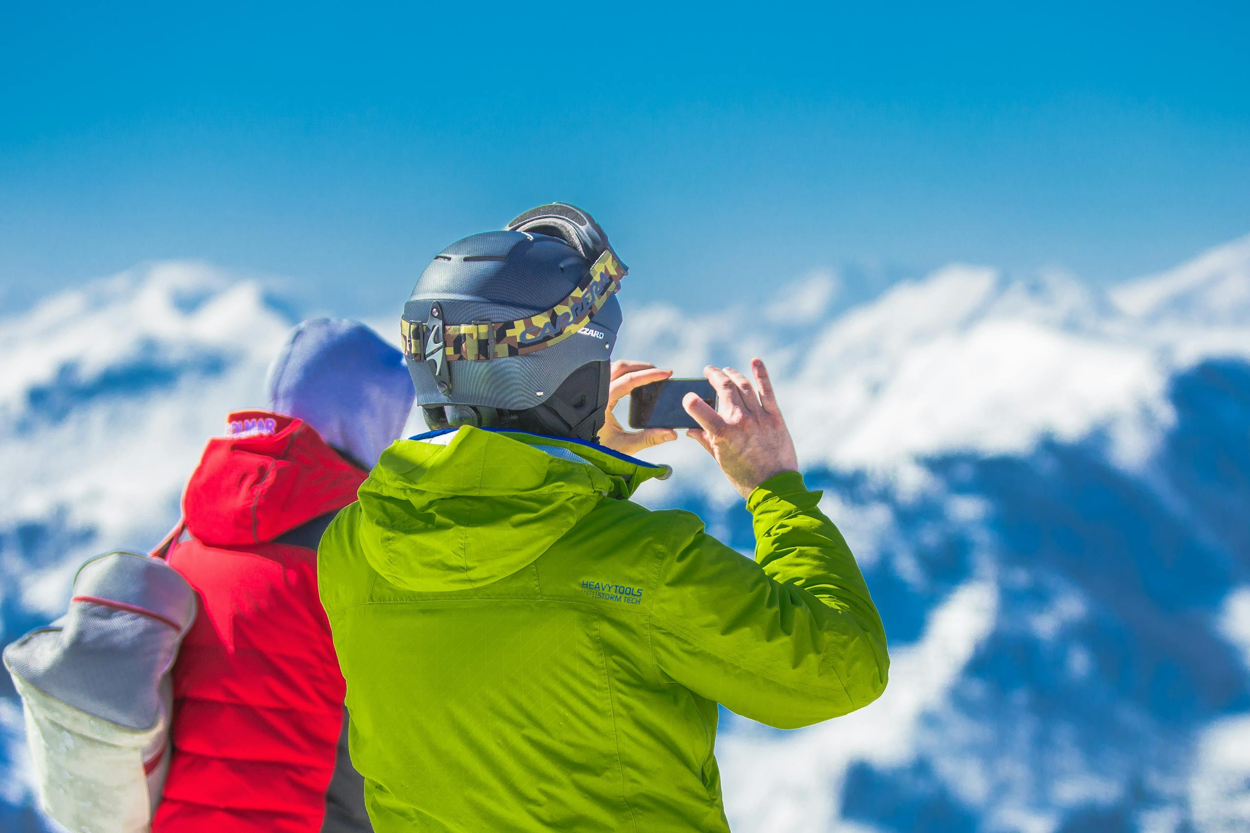 Man in Green Jacket and Gray Helmet Holding Phone Standing Next to Person in Red and Black Jacket