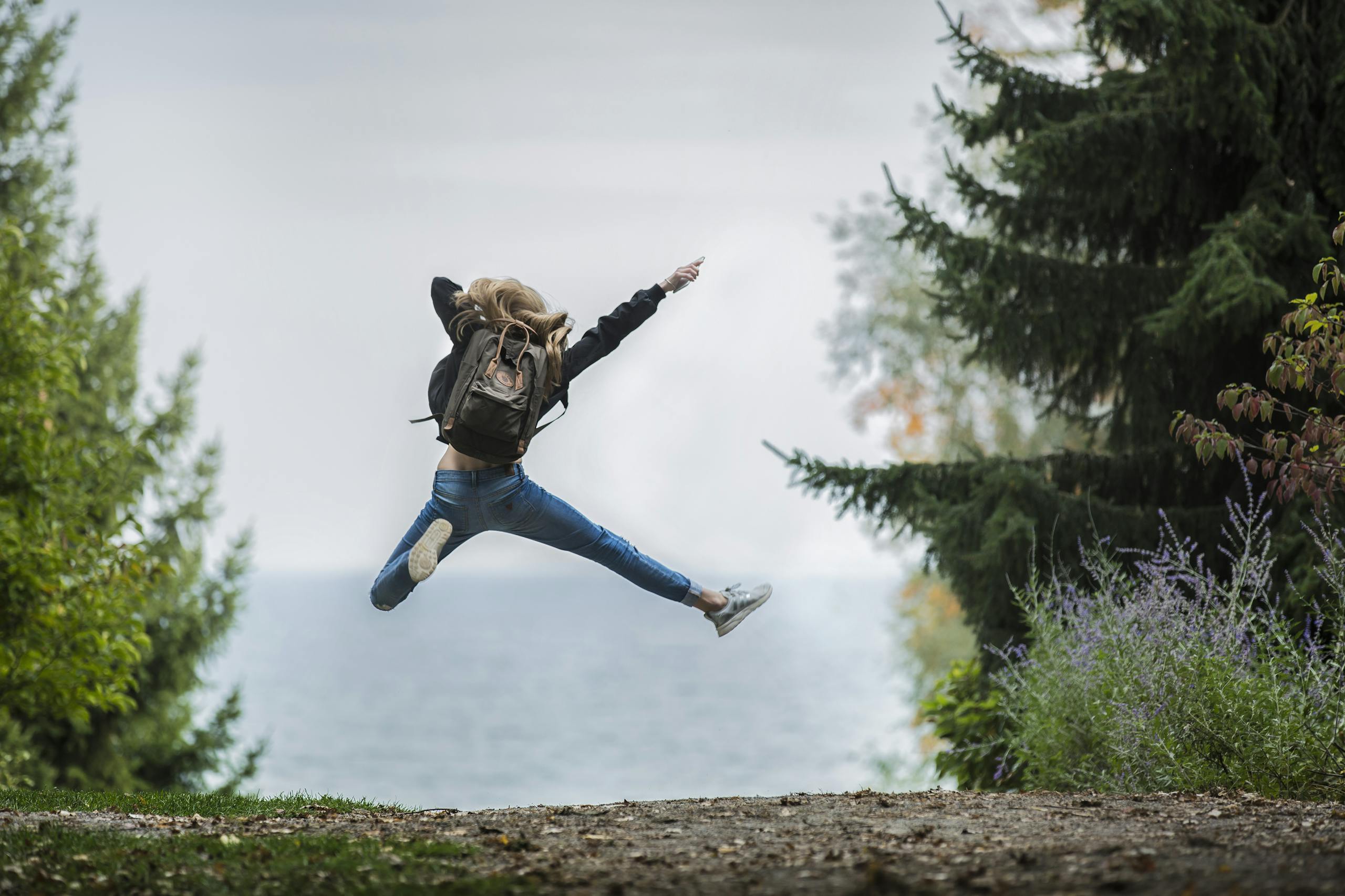 Energetic woman leaping in outdoor forest setting with lake view, showcasing freedom and joy.